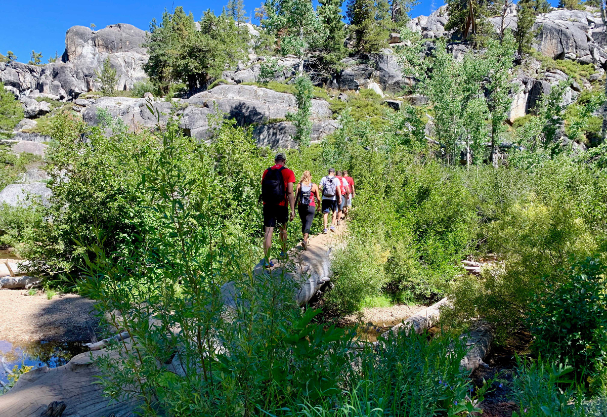 Hikers cross a stream on the trail to Lake Margaret on a log that serves as a bridge.