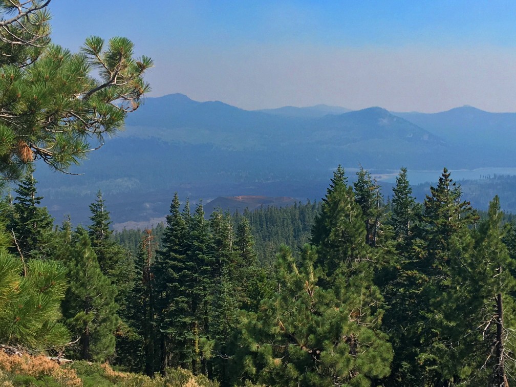 The top of Cinder Cone can be seen just barely above the treeline through the smokey haze with the Fantastic Lava Beds behind it and Butte Lake behind the beds.