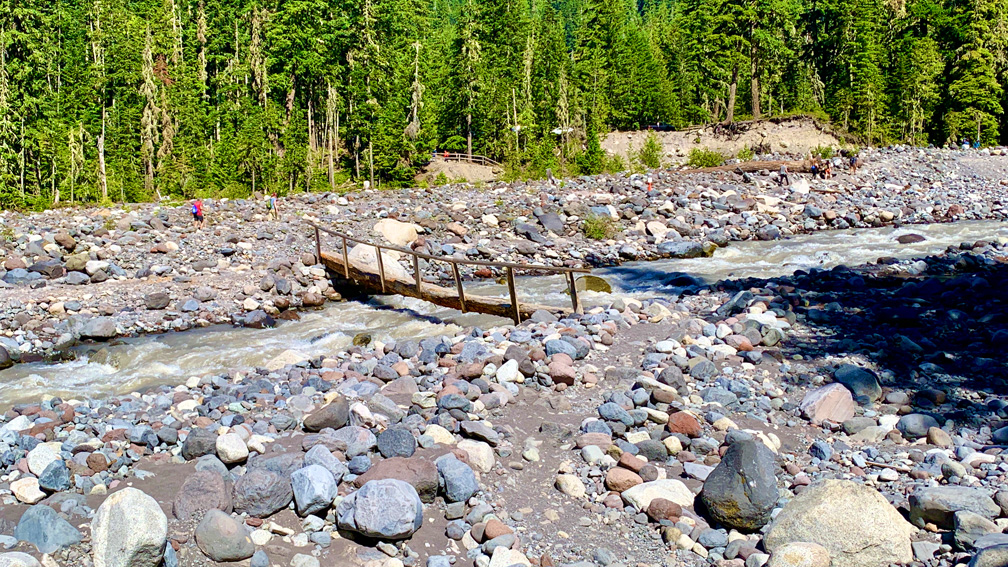 A log with a wooden railing attached to it crosses the fast moving Nisqually River on the way to Carter Falls.