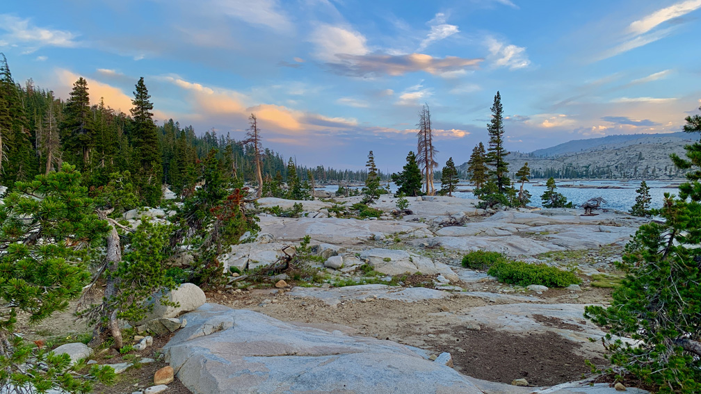 Granite rocks line the shore of the Lake Aloha, which is behind sparse pine trees.